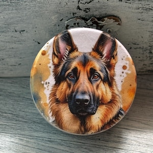 May include: A round, white button with a colorful image of a German Shepherd dog. The dog's fur is brown and black, and its eyes are brown. The background is a watercolor-style design with splashes of yellow, orange, and blue.