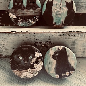 May include: Four round decorative magnets featuring black cats. One magnet shows two kittens peering over a wooden fence. Another shows two cats silhouetted by a window. The other two magnets show close-ups of a black cat's face.