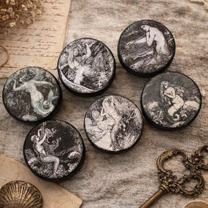 May include: A collection of six round, black and white decorative items featuring mermaid illustrations. The items are arranged on a textured, neutral-toned surface, alongside a seashell and a vintage key. The illustrations depict mermaids in various poses.