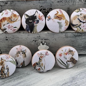 May include: A collection of eight round decorative magnets featuring watercolor illustrations of cats. The cats are depicted in various poses, interacting with butterflies and flowers. The color palette includes shades of orange, black, white, and pink.