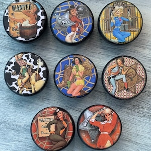May include: Set of seven round, black-rimmed coasters with colorful images of pin-up style women in various western-themed scenes. The coasters feature images of women in cowboy hats, riding horses, and holding guns. The text "WANTED" and "SHERIFF" are visible on some of the coasters.