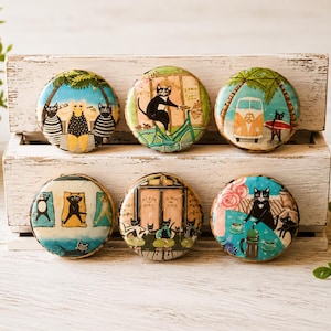 May include: Six circular magnets featuring colorful illustrations of black and white cats in various scenes, including beach relaxation, cycling, surfing, yoga, dining, and coffee time, arranged in two rows on a light-colored wooden crate.