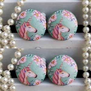 May include: Four round earrings with a unicorn design. Each earring features a unicorn with a pink mane and a horn, surrounded by pink flowers and green leaves on a light green background. The earrings are set against a white background.