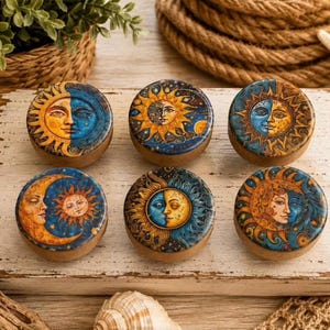 May include: A set of six round wooden knobs, each featuring a unique celestial design. The tops display detailed illustrations of suns, moons, and stars in vibrant shades of blue, gold, and orange, set against a natural light wood base.