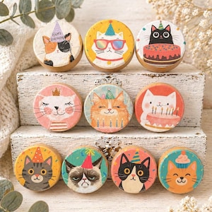 May include: A collection of ten round wooden cat-themed decorations. Each decoration features a different cat illustration, some wearing party hats or sunglasses, and some with birthday cakes. The color palette includes orange, yellow, black, and white.