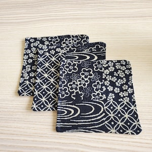 May include: Three square coasters with a navy blue background and white floral and geometric patterns. The coasters are made of fabric and have rounded corners. The patterns include flowers, circles, and water-like designs.