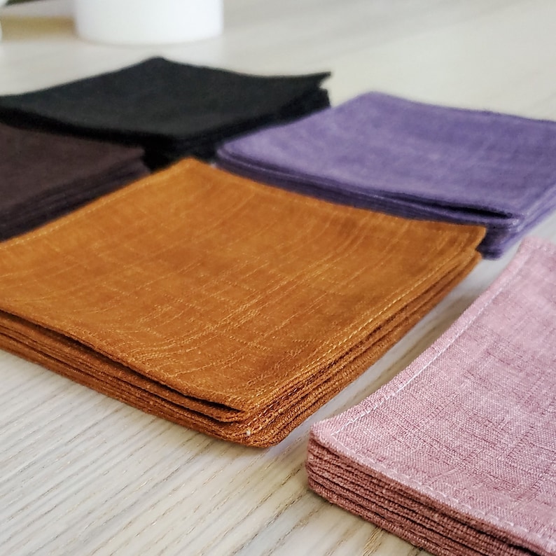 May include: A stack of four different colored linen napkins. The napkins are folded in a square shape and are arranged in a row. The colors are black, purple, brown, and pink.