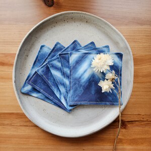 May include: A stack of six blue and white tie-dye coasters with a stitched edge. The coasters are arranged in a circle on a light gray ceramic plate. A dried flower is placed on top of the coasters.