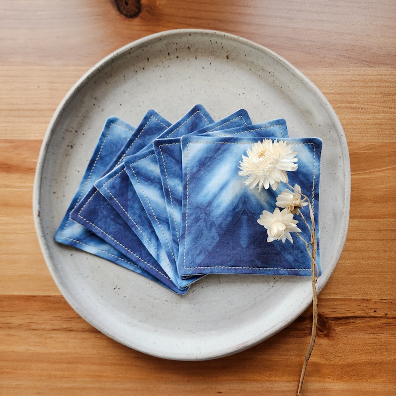 May include: A stack of six blue and white tie-dye coasters with a stitched edge. The coasters are arranged in a circle on a light gray ceramic plate. A dried flower is placed on top of the coasters.