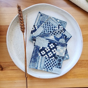 May include: Four blue and white fabric coasters with a patchwork design. The coasters are on a white ceramic plate with a speckled finish.