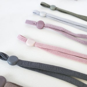 May include: Assortment of five different colored elastic cords with adjustable toggles. The cords are in shades of gray, pink, and light purple.