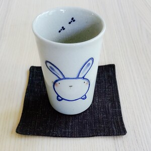 May include: A white ceramic teacup with a blue bunny illustration. The cup is sitting on a black fabric coaster.