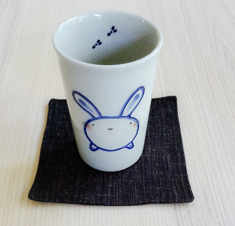 May include: A white ceramic teacup with a blue bunny illustration. The cup is sitting on a black fabric coaster.