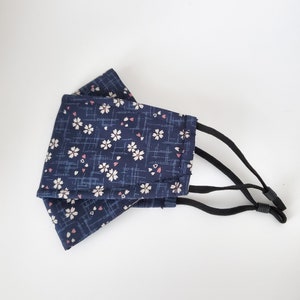 May include: A dark blue face mask with a white floral pattern and pink accents. The mask has adjustable black elastic straps.
