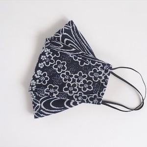以下が含まれることがあります： 白い花の模様が入った濃い青色のマスク。マスクにはゴム製の耳ひもが付いています。