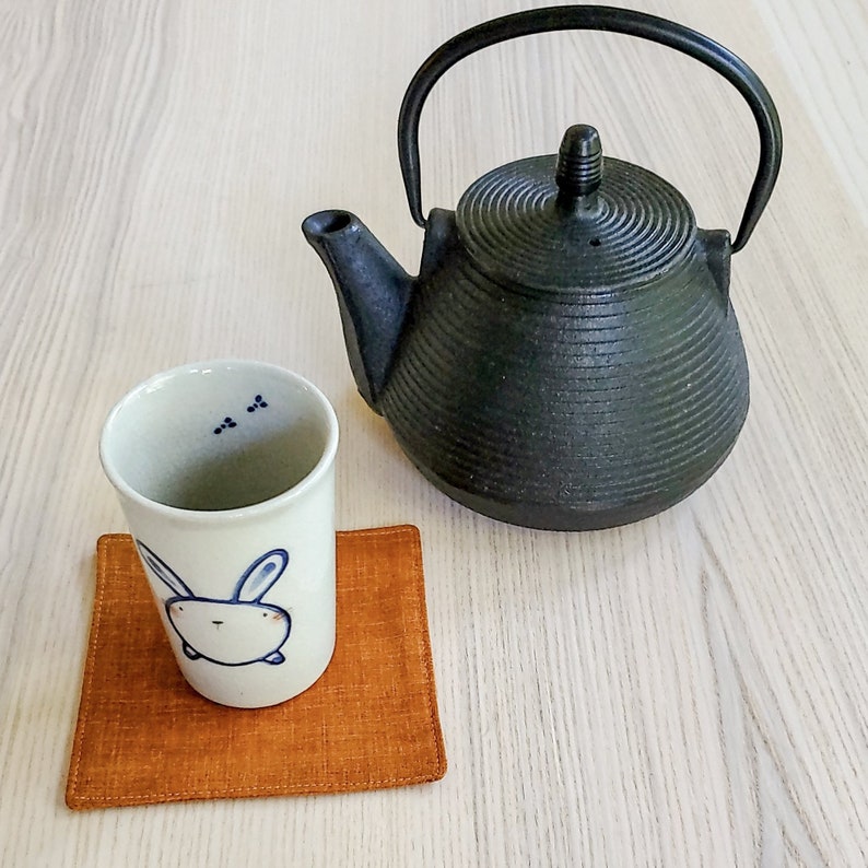 May include: A white ceramic teacup with a blue bunny design sits on a brown coaster. A black cast iron teapot with a rounded body and a black handle is on a light wood surface.