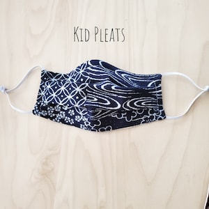 Puede incluir: Una mascarilla facial plisada de color negro y blanco con elásticos blancos. La mascarilla tiene un diseño plisado y el texto "Kid Pleats" está impreso encima de la mascarilla.