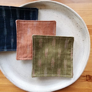 May include: Three square fabric coasters in different patterns. The coasters are on a white ceramic plate. The coasters are made of a woven fabric with a subtle pattern. The coasters are in shades of blue, orange, and green.