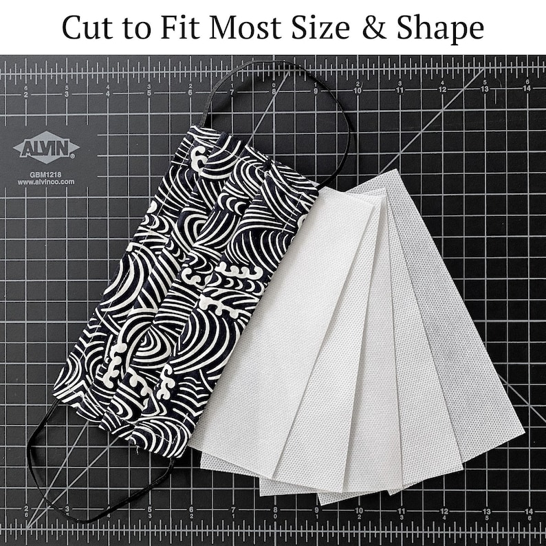 May include: A black and white patterned fabric face mask with a black elastic band. The mask is laid out on a black cutting mat with five white filter inserts.