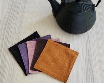 May include: Five fabric coasters in a variety of colours, including black, purple, pink, brown, and orange. The coasters are arranged in a stack, with the black coaster at the bottom and the orange coaster at the top. The coasters are made from a soft, textured fabric.