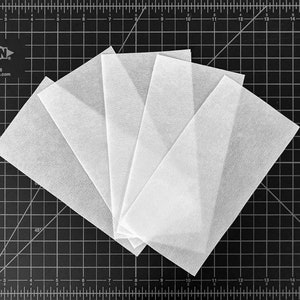 May include: Five white fabric squares arranged in a fan shape on a black cutting mat with a grid pattern. The mat has inch markings.