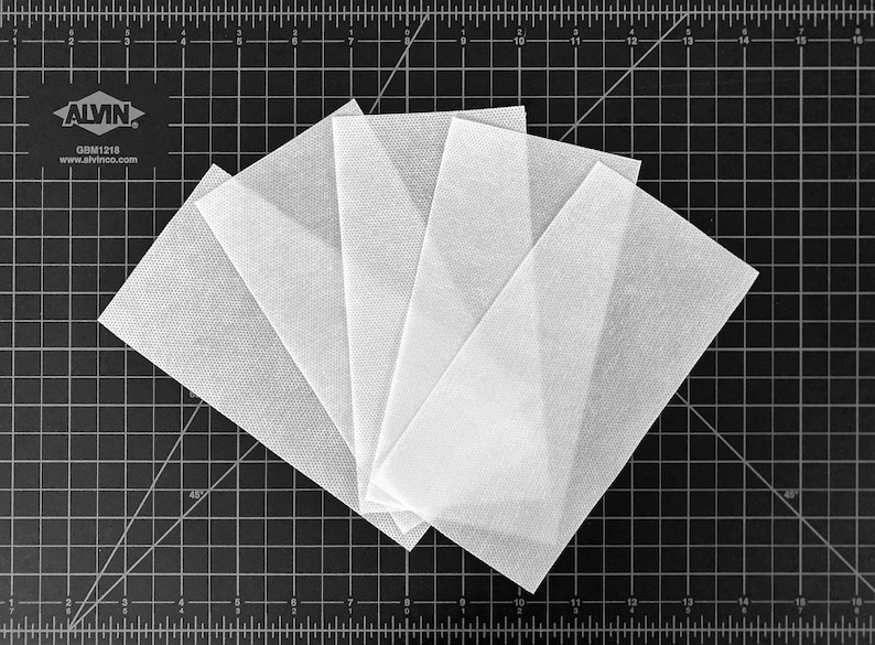 May include: Five white fabric squares arranged in a fan shape on a black cutting mat with a grid pattern. The mat has inch markings.