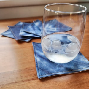 May include: A glass of water with ice cubes sitting on a blue and white tie-dye coaster. The coaster has a yellow stitched border.
