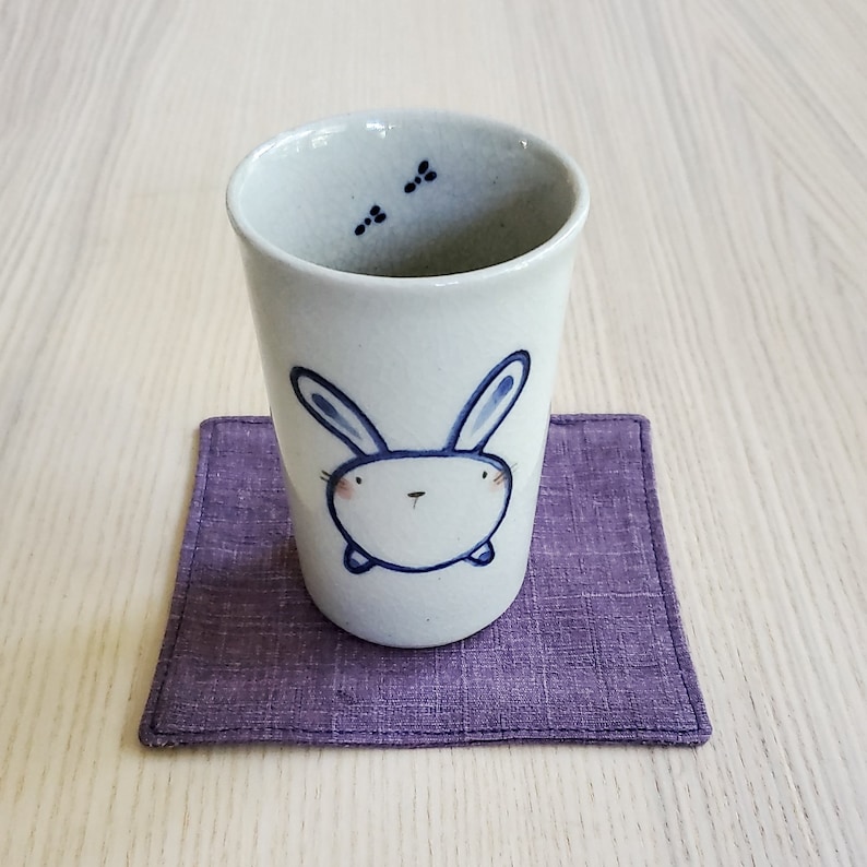 May include: A white ceramic teacup with a blue bunny design on the front. The cup is sitting on a purple fabric coaster.