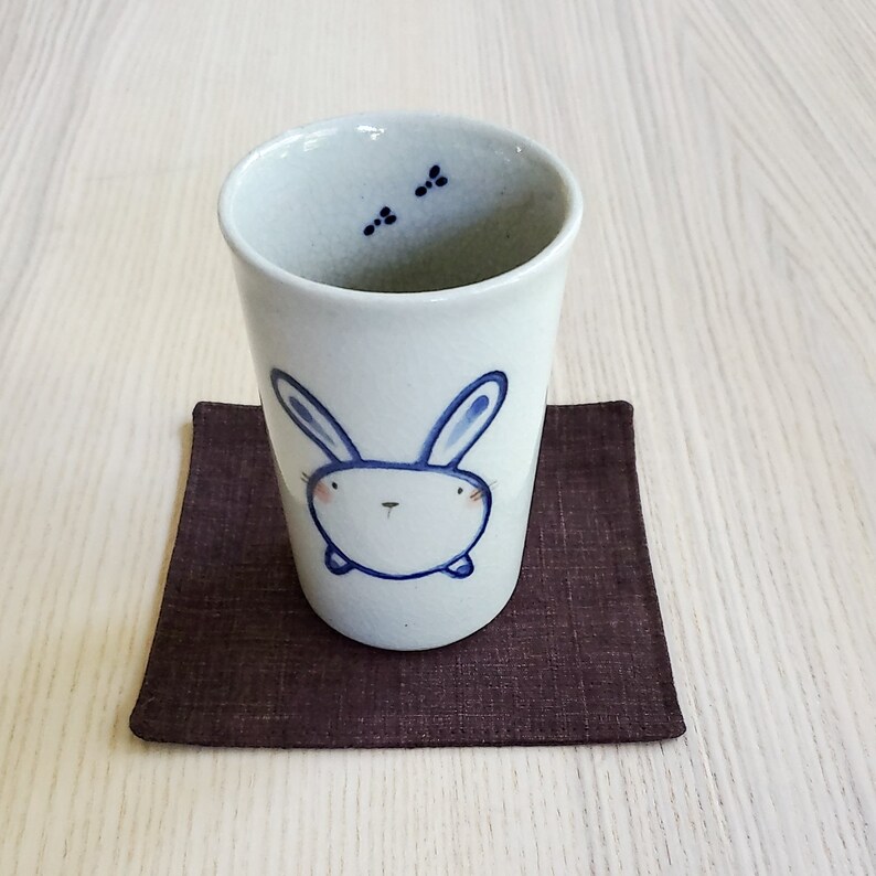 May include: A white ceramic mug with a blue bunny illustration. The mug is sitting on a brown coaster.