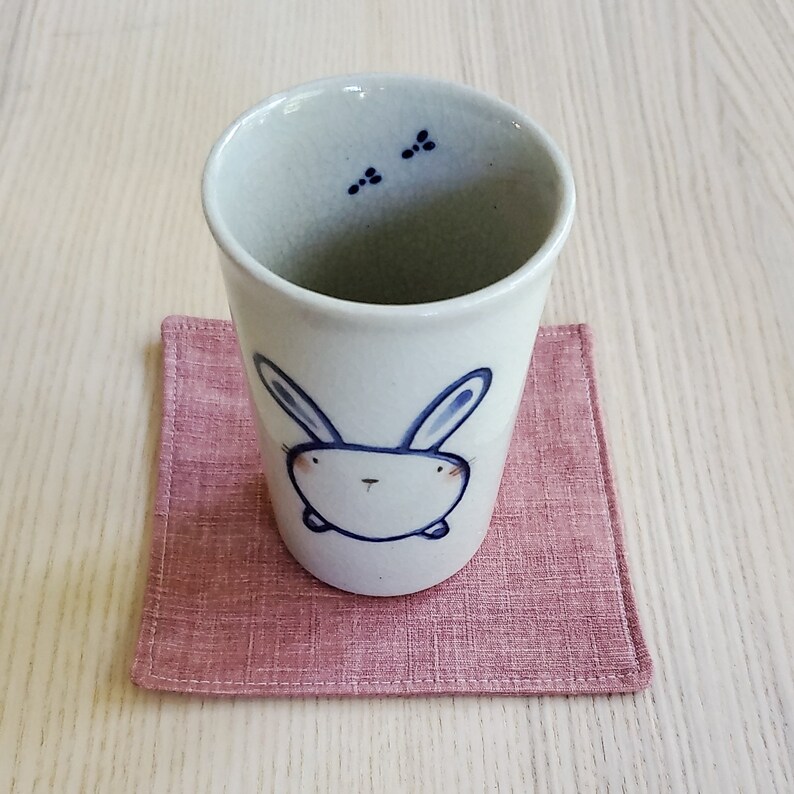 May include: A white ceramic mug with a blue bunny illustration on the front. The mug is sitting on a pink linen coaster.