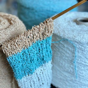 May include: Close-up of a crocheted fabric with three stripes of beige, turquoise, and white. The fabric is being worked on with a wooden knitting needle.
