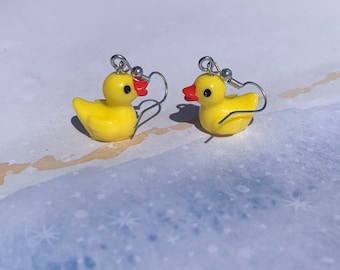 May include: A pair of whimsical yellow duck earrings with red beaks and black eyes. The earrings are made of a glossy material and feature silver-colored ear wires. These novelty earrings are a fun accessory.