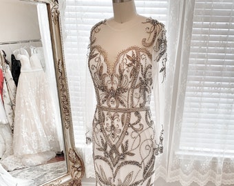 May include: A white beaded gown with a sheer bodice and long sleeves. The gown features intricate floral designs and beading throughout.
