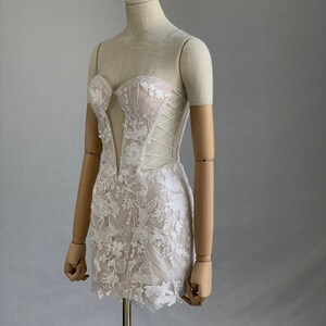 May include: A white lace mini dress with a sweetheart neckline and a fitted bodice. The dress has a lace overlay with floral embroidery and a corset-style lacing detail in the back. The dress is on a mannequin wearing a silver tiara.