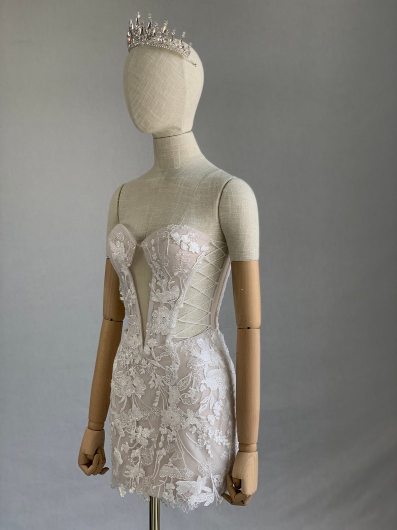 May include: A white lace mini dress with a sweetheart neckline and a fitted bodice. The dress has a lace overlay with floral embroidery and a corset-style lacing detail in the back. The dress is on a mannequin wearing a silver tiara.