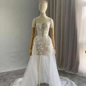 May include: A white strapless wedding dress with a lace bodice and a tulle skirt. The dress has a long train and is decorated with delicate floral embroidery. The dress is displayed on a mannequin wearing a tiara.