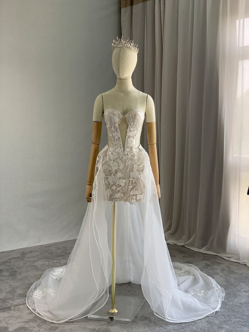 May include: A white strapless wedding dress with a lace bodice and a tulle skirt. The dress has a long train and is decorated with delicate floral embroidery. The dress is displayed on a mannequin wearing a tiara.