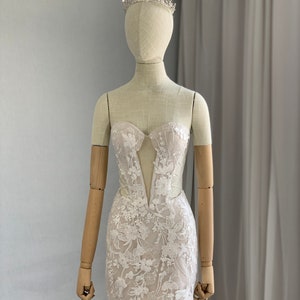 May include: A white lace mini dress with a plunging neckline and a fitted silhouette. The dress is adorned with delicate floral embroidery and has a scalloped hem. The dress is being displayed on a mannequin wearing a silver tiara.
