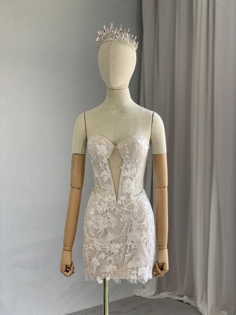 May include: A white lace mini dress with a plunging neckline and a fitted silhouette. The dress is adorned with delicate floral embroidery and has a scalloped hem. The dress is being displayed on a mannequin wearing a silver tiara.