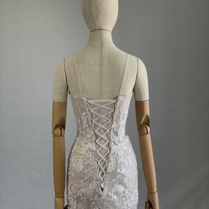 May include: A white lace mini dress with a corset back and floral embroidery. The dress is on a mannequin with wooden arms.