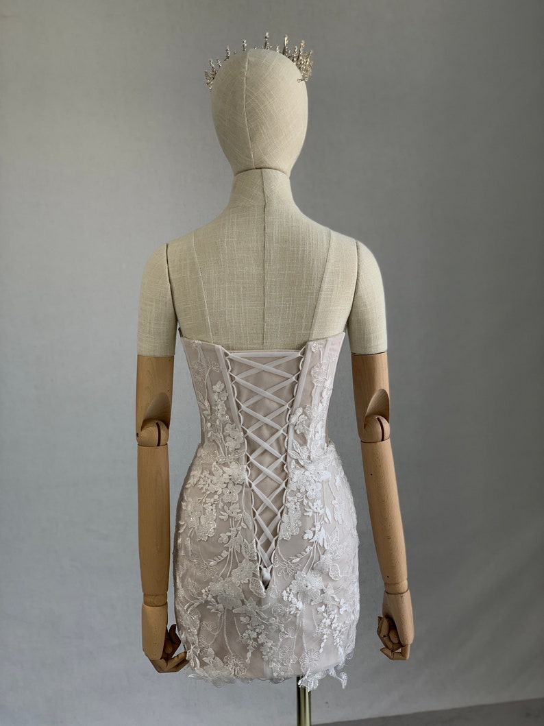 May include: A white lace mini dress with a corset back and floral embroidery. The dress is on a mannequin with wooden arms.