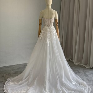 May include: A white tulle wedding dress with a lace bodice and a long train. The dress has a sweetheart neckline and a fitted bodice with a lace-up back. The skirt is full and flowing, and the dress is adorned with delicate lace appliques.