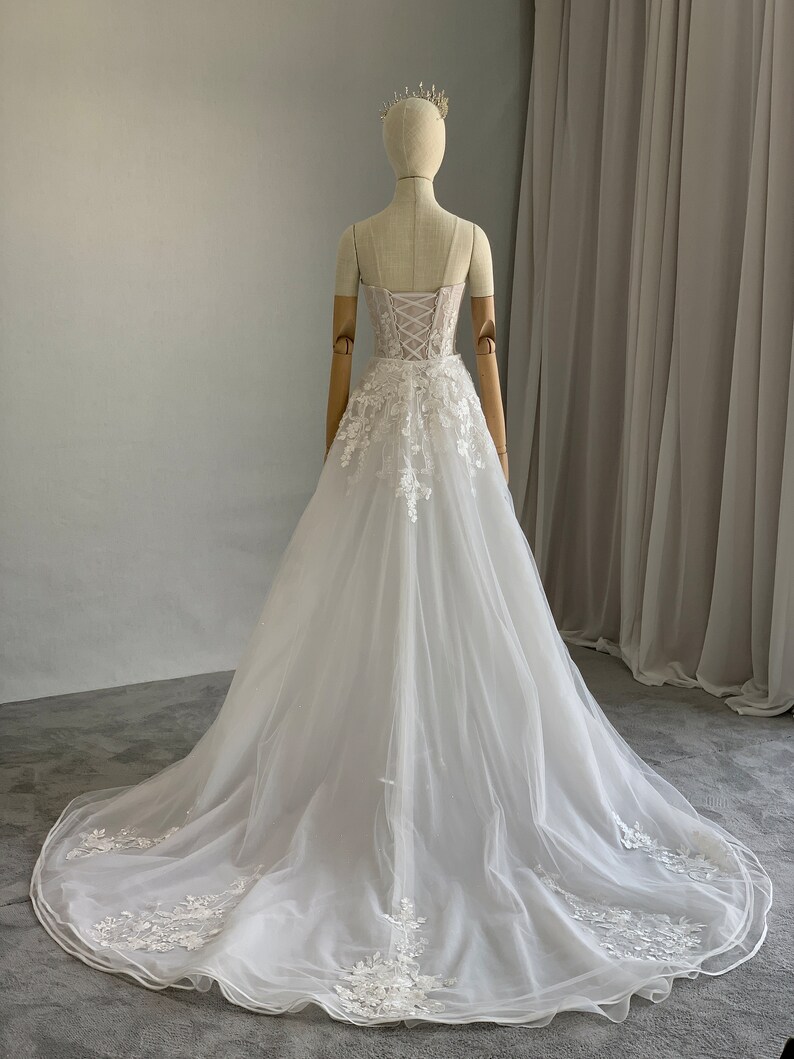 May include: A white tulle wedding dress with a lace bodice and a long train. The dress has a sweetheart neckline and a fitted bodice with a lace-up back. The skirt is full and flowing, and the dress is adorned with delicate lace appliques.