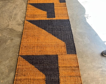 May include: A rectangular runner rug with a geometric design. The rug features a pattern of dark navy blue shapes against a background of golden-orange woven material. The rug is placed on a concrete floor.