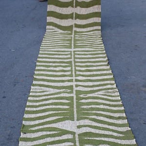 May include: A long, rectangular rug with a green and white striped pattern. The rug has a central vertical stripe and a fringe at the bottom. The text "MAJESTICRUGDECOR" is visible at the bottom. The rug is displayed outdoors.
