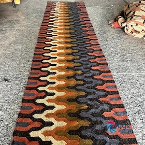 Puede incluir: Una alfombra rectangular con un patrón geométrico en tonos naranja, marrón, negro y azul. La alfombra tiene un borde con flecos y un diseño repetitivo. La alfombra está hecha de material tejido.