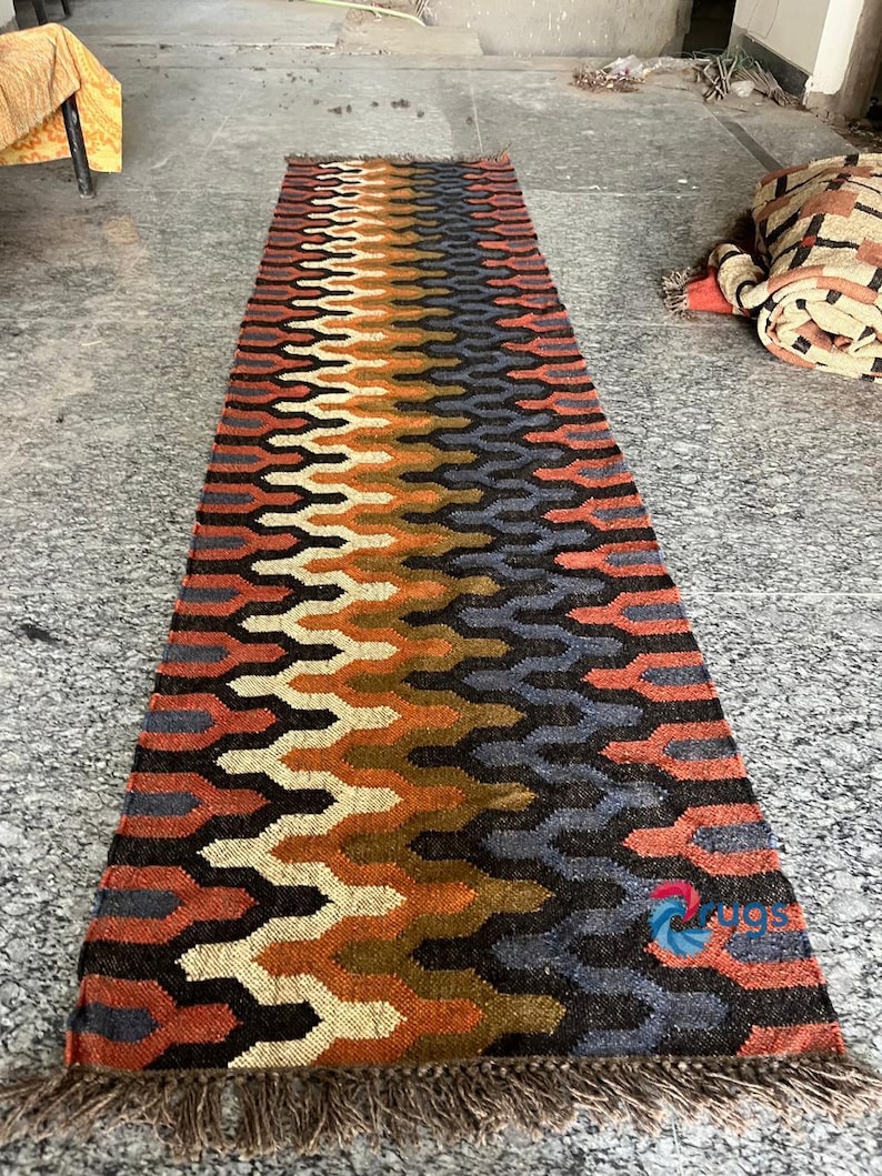 Puede incluir: Una alfombra rectangular con un patr&oacute;n geom&eacute;trico en tonos naranja, marr&oacute;n, negro y azul. La alfombra tiene un borde con flecos y un dise&ntilde;o repetitivo. La alfombra est&aacute; hecha de material tejido.
