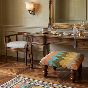 Pode incluir: Um conjunto de toucador vintage com uma secretária de madeira, cadeira e banco. A secretária tem gavetas e é adornada com frascos, pincéis e uma caixa de joias. O banco apresenta um padrão geométrico colorido. A sala tem uma estética clássica e elegante.