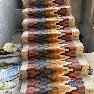 Puede incluir: Una alfombra de pasillo para escaleras con un patr&oacute;n geom&eacute;trico en tonos naranja, marr&oacute;n, gris y azul. La alfombra est&aacute; hecha de material tejido y tiene un borde con flecos. Se coloca sobre escalones de ladrillo.