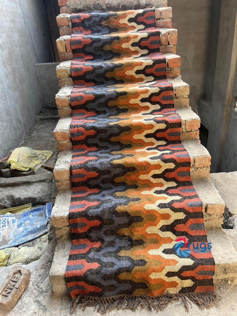 Puede incluir: Una alfombra de pasillo para escaleras con un patr&oacute;n geom&eacute;trico en tonos naranja, marr&oacute;n, gris y azul. La alfombra est&aacute; hecha de material tejido y tiene un borde con flecos. Se coloca sobre escalones de ladrillo.